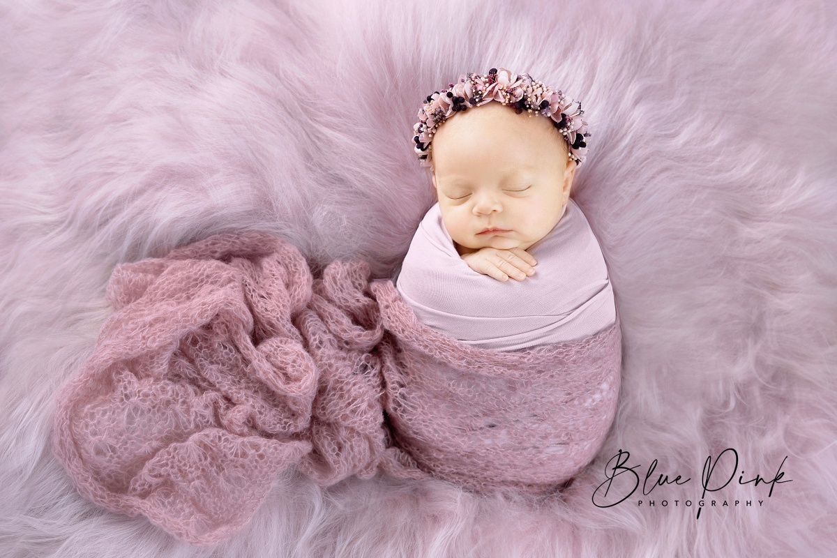  Newborn baby girl in a serene pose, wrapped in a soft pale pink knitted wrap with a delicate flower halo. Closed eyes and tiny hands outside the wrap create a precious and heartwarming portrait for a memorable newborn photography session.